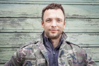 Young Caucasian man in camouflage. Outdoor portrait over green rural wooden wall