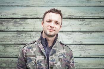 Smiling young Caucasian man in camouflage. Outdoor portrait over green rural wooden wall