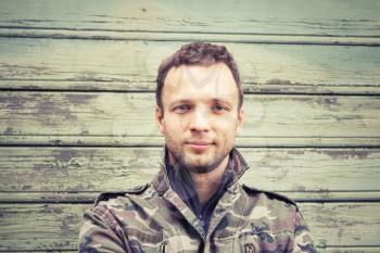 Young Caucasian man in camouflage. Outdoor portrait over green rural wooden wall. Vintage warm tonal photo filter effect