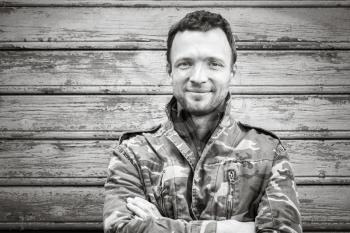 Young smiling Caucasian man in camouflage, outdoor black and white portrait over rural wooden wall