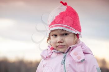 Cute Caucasian blond baby girl in pink hat angry frowns, outdoor closeup portrait 