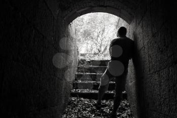 Young man stands in dark stone tunnel with glowing end, black and white photo