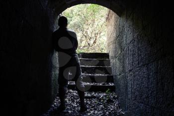 Young man stands in dark stone tunnel with glowing end, vintage tonal correction filter effect