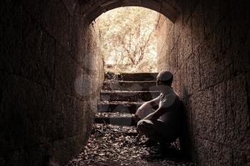 Young man sits inside of dark stone tunnel with glowing end, warm tonal correction photo filter