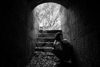 Young sad man sitting inside of dark stone tunnel with stairs in the end, black and white photo