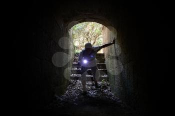 Young man with a flashlight enters the stone tunnel and looks inside with attention and fear
