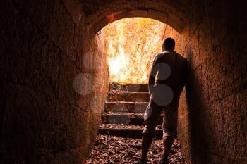 Young man stands in dark stone tunnel with big fire in the end