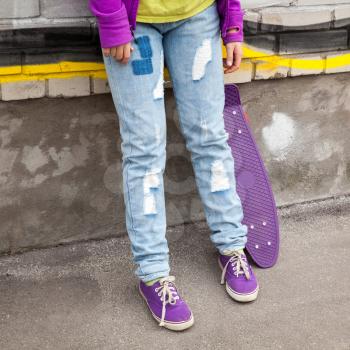 Teenager in blue jeans and gumshoes stands with skateboard near urban wall