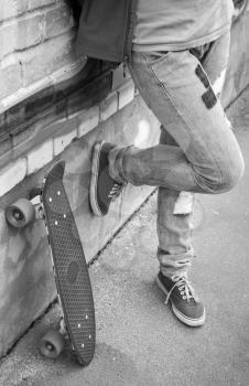Teenager in jeans and gumshoes stands with skateboard near brick wall