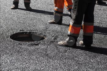 Urban road under construction, asphalting in progress, workers near sewer manhole, feet fragment