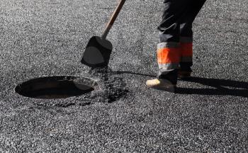 Urban road under construction, asphalting in progress, worker with a shovel near sewer manhole, feet fragment