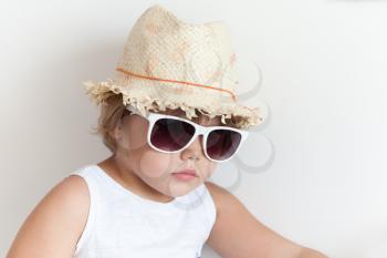 Cute Caucasian little girl in straw hat and sunglasses on white wall background