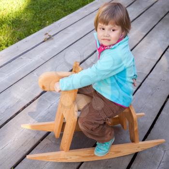 Outdoor portrait of cute Caucasian blond baby girl riding small wooden horse toy