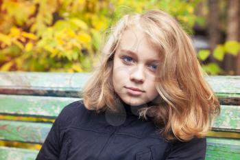 Beautiful Caucasian blond teenage girl in black jacket sitting on old green bench in autumn park