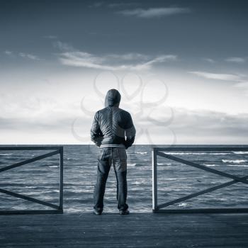 Young man looking on the sea from wooden pier. Rear view, blue toned monochrome square photo