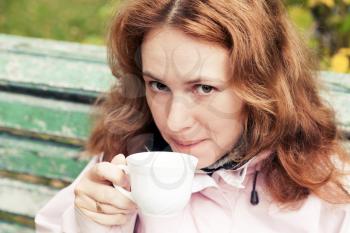 Young adult Caucasian woman drinks coffee from white cup in autumnal park