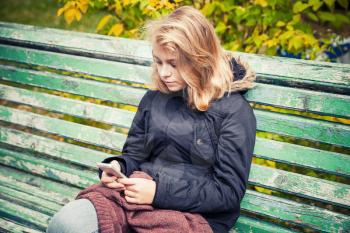 Beautiful Caucasian blond teenage girl in black jacket sitting on old green park bench with smart phone in hands