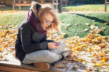 Cute Caucasian blond teenage girl in jeans, scarf and black jacket sitting on park bench and using smartphone for messaging, outdoor autumn portrait