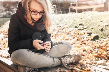 Caucasian blond teenage girl in jeans and black jacket sitting on wooden park bench and using smartphone, outdoor autumn portrait, vintage style tonal correction photo filter