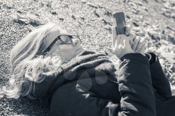Caucasian blond teenage girl in black jacket laying in park and using cellphone, monochrome photo