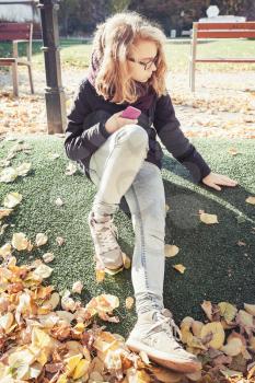 Beautiful blond teenage girl in glasses sitting in autumnal European city park with falling leaves, vintage style tonal correction photo filter