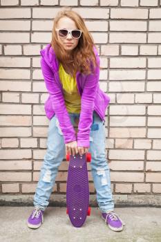 Blond teenage girl in jeans, sunglasses and sporty clothes holds skateboard near by gray urban brick wall