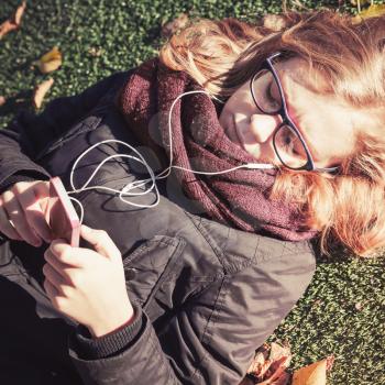 Caucasian blond teenage girl laying in autumnal park and using smart phone with headphones, vintage style tonal correction filter, square photo