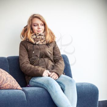 Beautiful blond Caucasian teenage girl in warm clothes sitting on blue sofa over white wall background