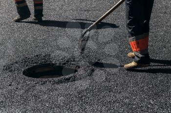 Urban road under construction, asphalting in progress, worker with shovel near sewer manhole, feet fragment