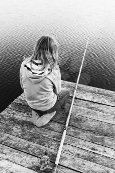 Little blond Caucasian girl sitting on a wooden pier with fishing rod, black and white photo, vertical composition