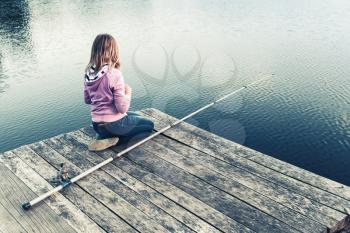 Little blond Caucasian girl sitting on a wooden pier with fishing rod, vintage stylized photo with tonal correction filter effect