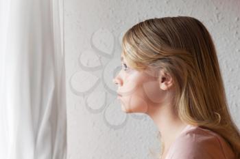 Beautiful blond Caucasian girl looking in a window with white curtains, close up portrait