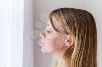 Closeup profile portrait of beautiful blond Caucasian girl with closed eyes near a window with white curtains