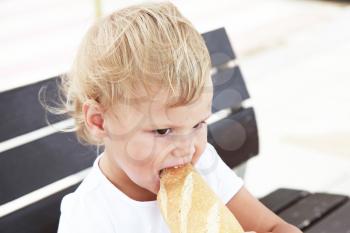 Outdoor close up portrait of cute Caucasian blond baby girl eating big French baguette