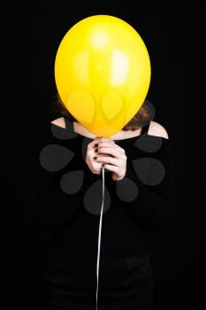 Girl hiding her face under yellow balloon, vertical studio shot over black background