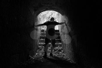 Young tired man leaves dark stone tunnel with glowing end, black and white photo 