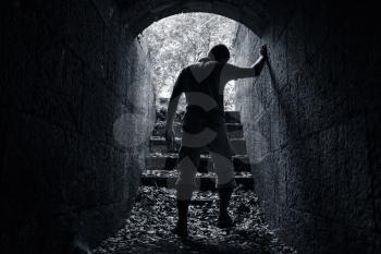 Young tired man leaves dark stone tunnel with glowing end, dark blue monochrome photo 