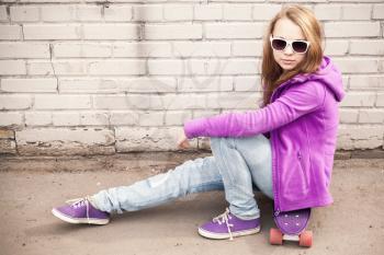 Blond teenage girl in jeans and sunglasses sits on skateboard near white brick wall, photo with warm retro tonal correction effect, old style filter
