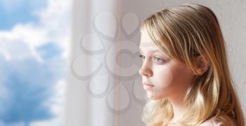 Closeup portrait of beautiful blond Caucasian girl, window with white curtains and cloudy blue sky outside are on a background