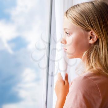 Closeup portrait of beautiful blond Caucasian girl standing near a window with white curtains and cloudy blue sky outside