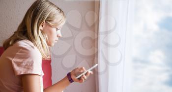 Cute Caucasian blond teenage girl in pink t-shirt using smartphone, profile portrait near open window