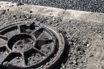 Urban road under construction, asphalting in progress. Dirty rusted sewer manhole cover lays on a roadside