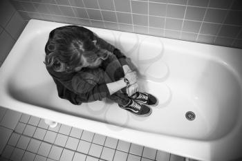 Depression concept. Stressed sad Caucasian teenage girl sitting in empty bath. Black and white photo