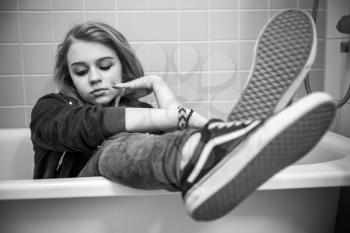 Teenage girl in black sporty sneakers sits in white bath. Black and white photo