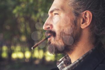 Bearded Asian man smoking cigar, outdoor profile portrait with selective focus and warm tonal correction filter effect