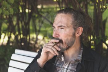 Bearded Asian man smoking cigar in summer park, outdoor portrait with selective focus