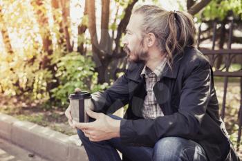 Bearded Asian man sitting on the sidewalk in the park with coffee in paper cup, outdoor portrait with selective focus and tonal correction effect
