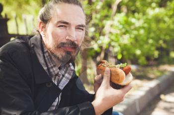 Bearded Asian man with hot dog has a lunch in summer park, outdoor portrait with selective focus