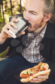 Bearded Asian man having lunch with hot dog and coffee in summer park, outdoor portrait with selective focus