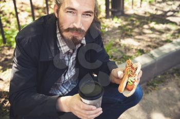 Bearded Asian man with hot dog and coffee in summer park, outdoor portrait with selective focus